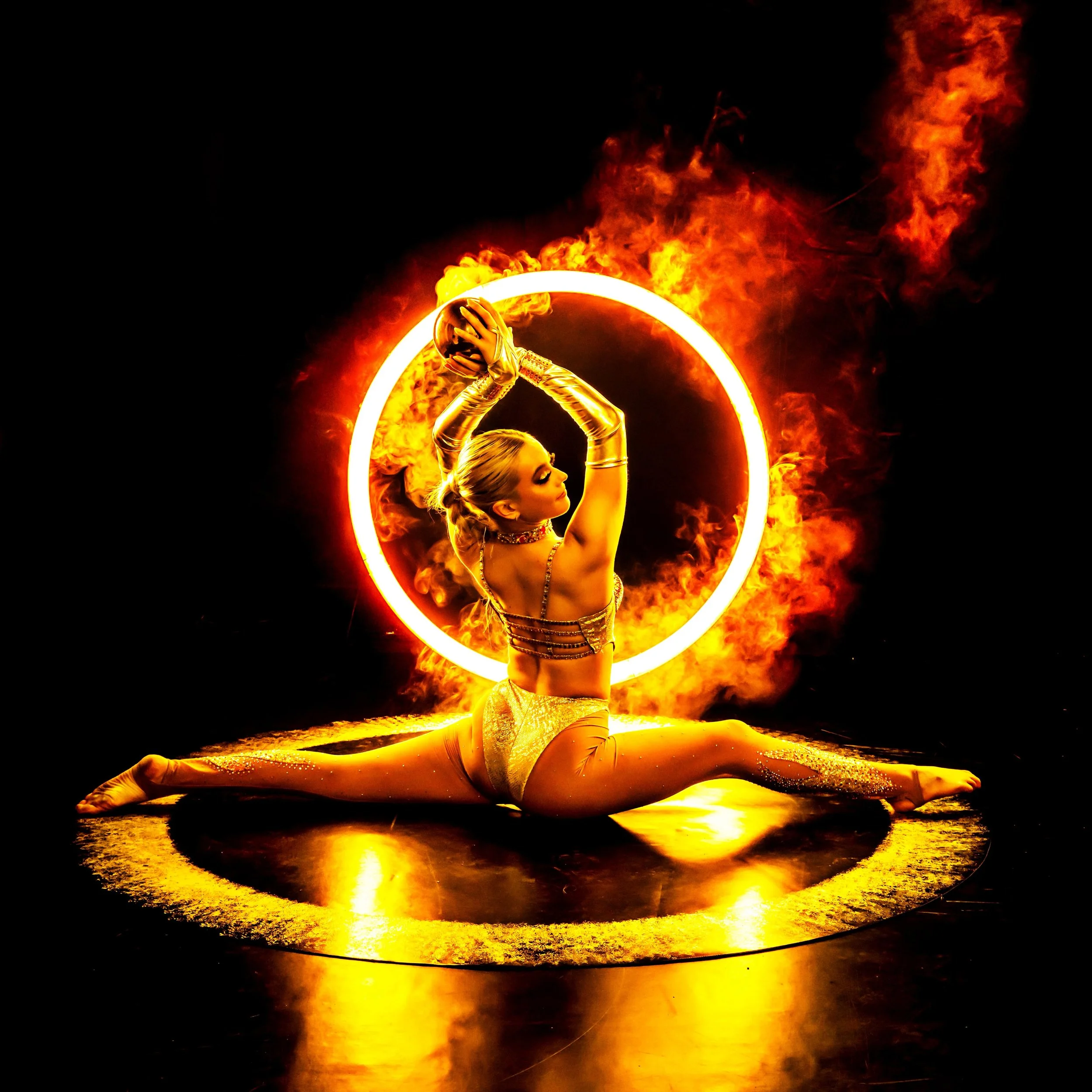 A female performer dressed in gold costume with long gloves, performing a fire dance while in the splits on stage, with fire rings around her and flames in the background.