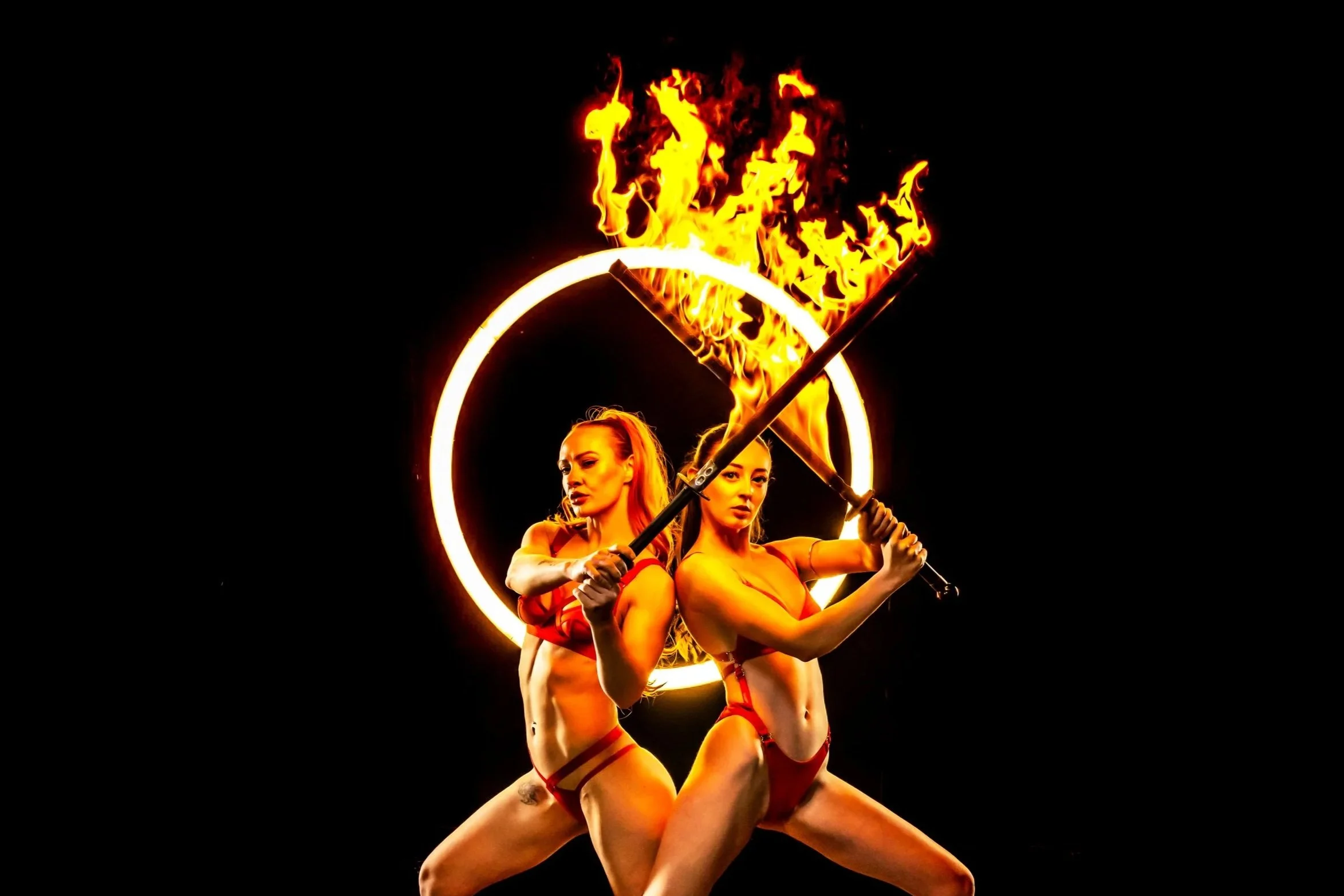 Two female fire dancers in red costumes performing at night, holding flaming fire props in front of a glowing circular frame.