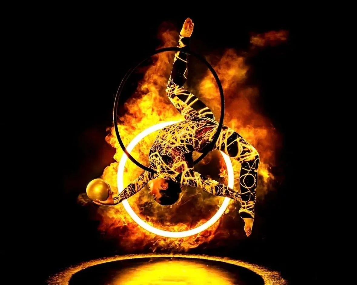 A performer dressed in a body suit with glowing patterns performs an acrobatic act on an aerial hoop amidst flames and fiery smoke, illuminated by bright circular lights.