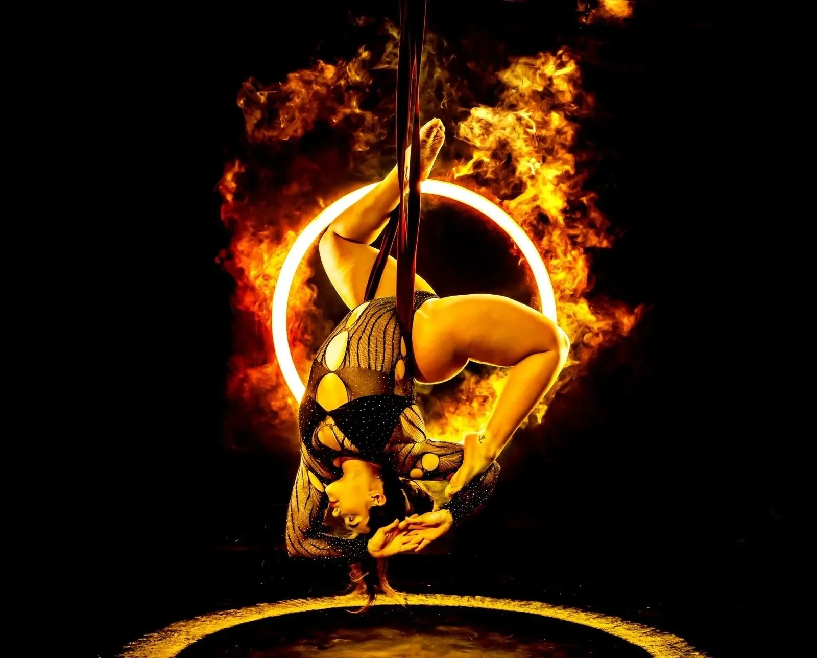 A female acrobat performing an aerial routine on a vertical silk, hanging upside down in front of flames and a glowing circle. She wears a black and yellow patterned costume against a dark background.