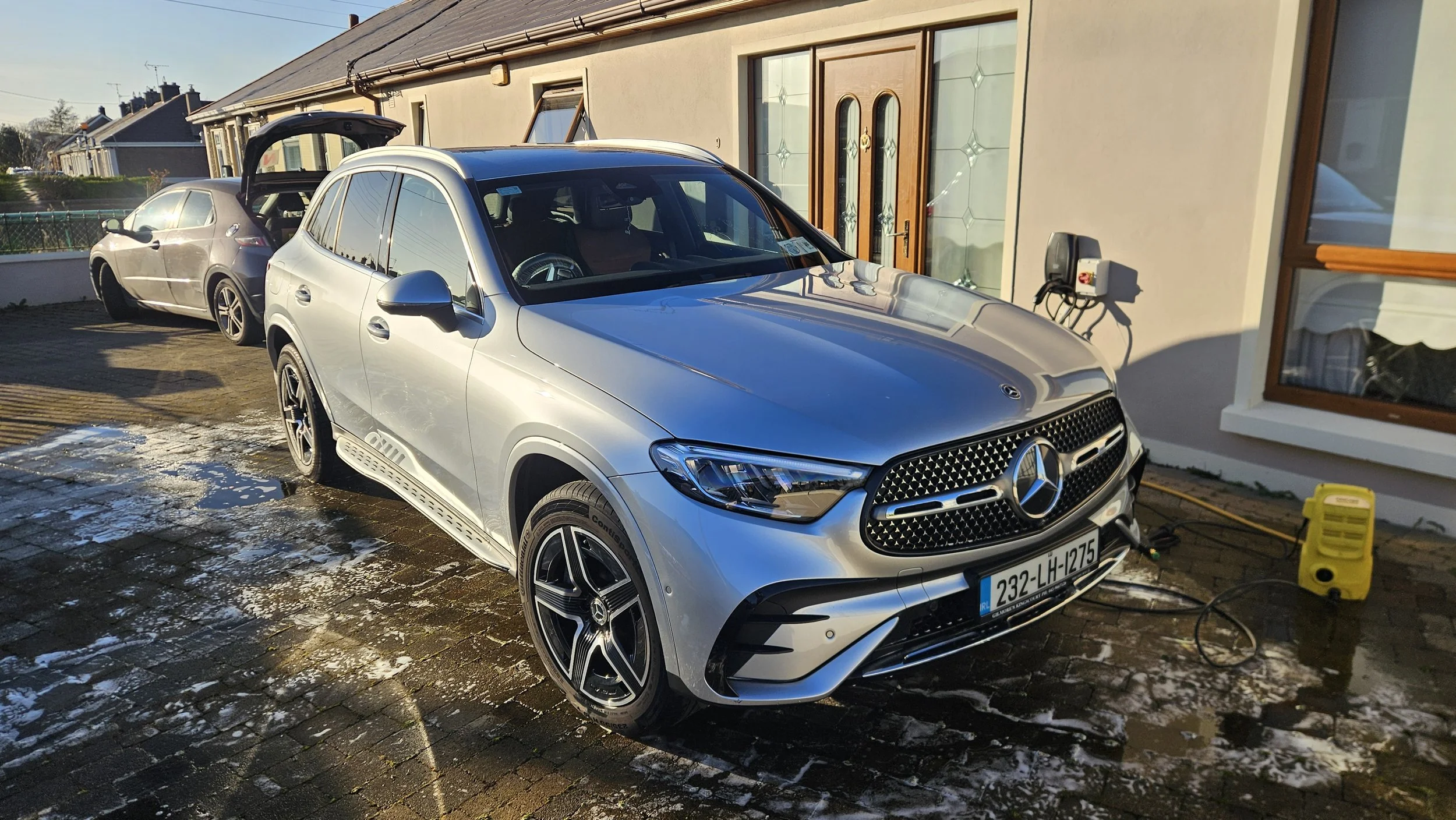 A silver Mercedes-Benz car is being washed or cleaned in a driveway next to a beige house with a wooden front door and large windows.