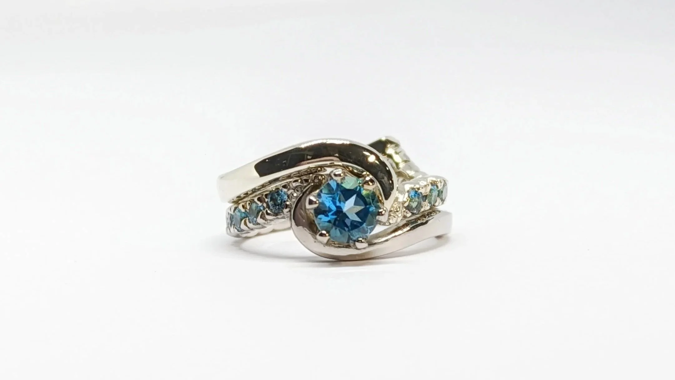 The engagement ring and wedding band interlock seamlessly, creating the appearance of a single, flowing piece. Crafted from heirloom white gold, the design features a striking London Blue Topaz, framed by a gradient of smaller accent topazes along th