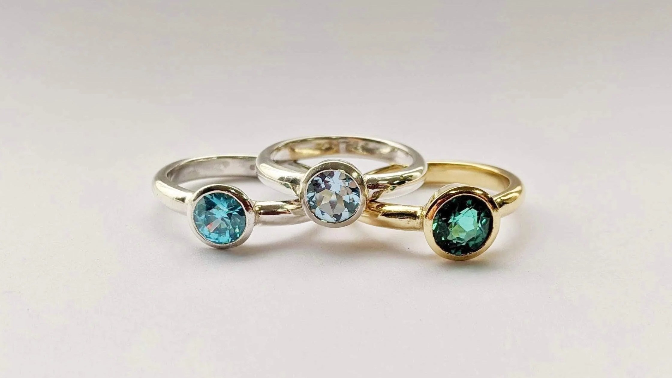 Trio. A set of three rings all with the same classic chunky solitaire design, but each with different materials and stones: 18ct yellow gold and an antique emerald, silver and a London blue topaz, silver and an aquamarine.