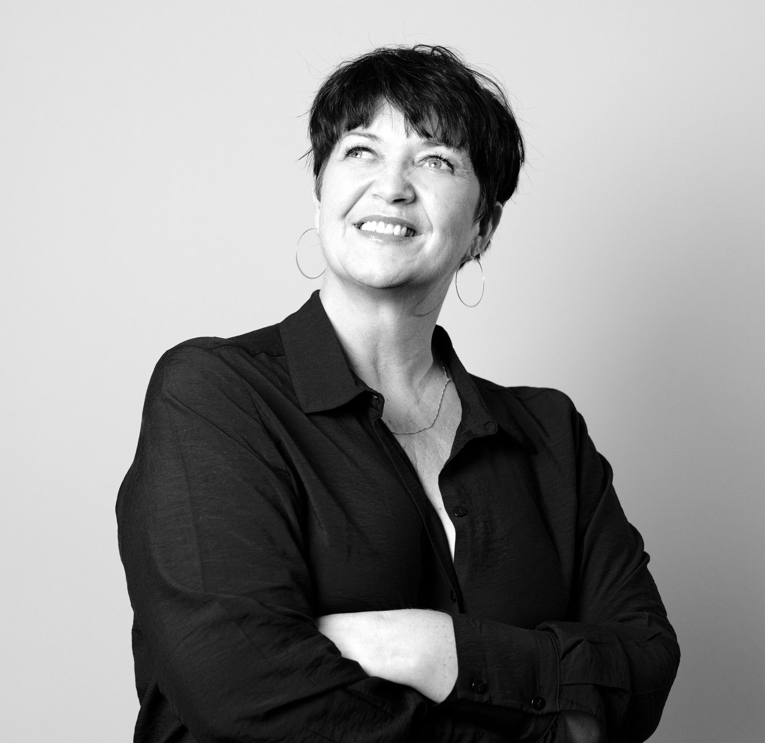 Sort-hvitt fotografi av en kvinne med kort mørkt hår som smiler og ser opp, iført svart jakke og ørepynt, mot en lys grå bakgrunn.