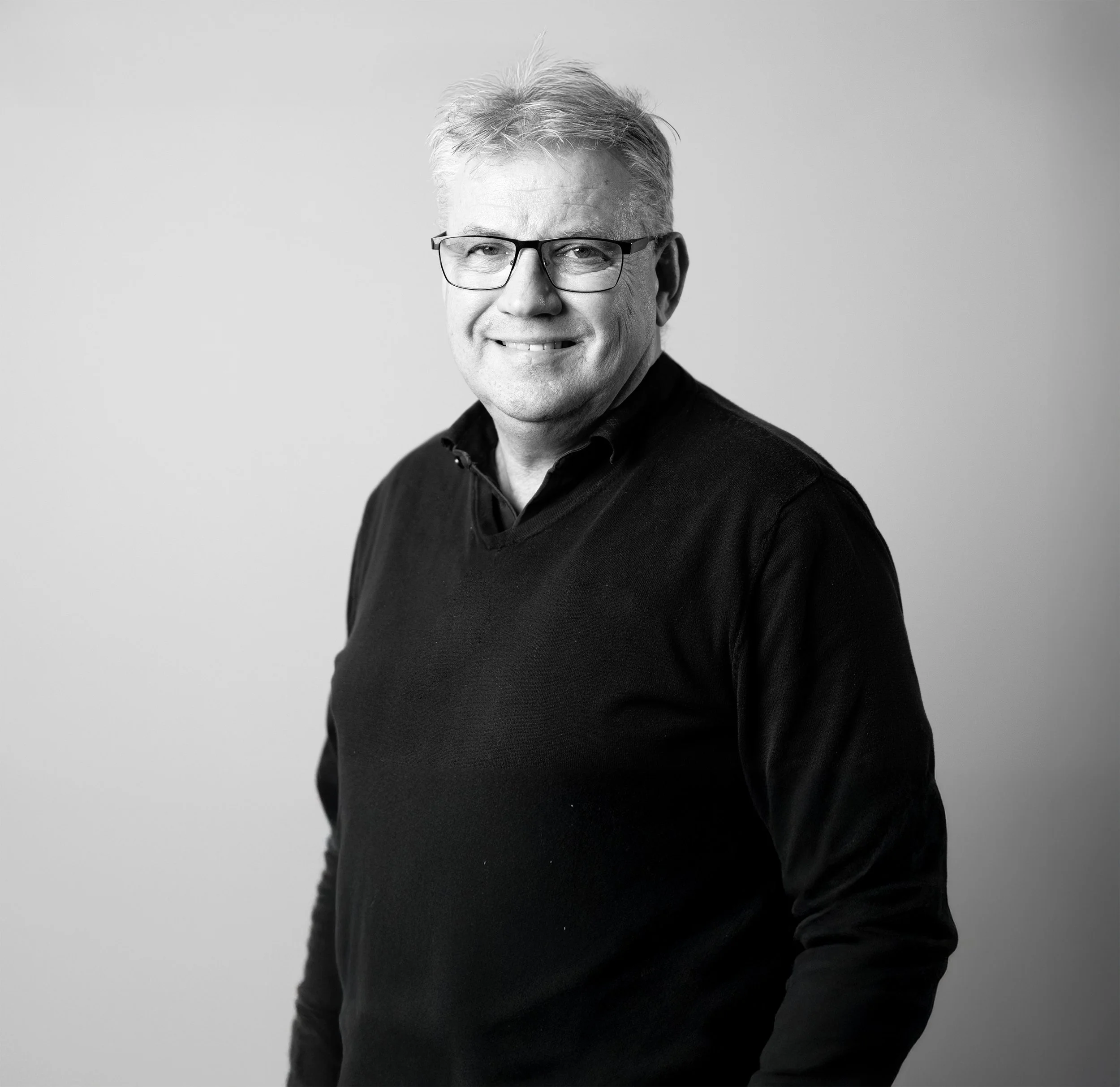 sort-hvitt fotografi av en mid-aged mann med grått hår, briller og svart genser, smilende på et nøytralt grått bakgrunn.
