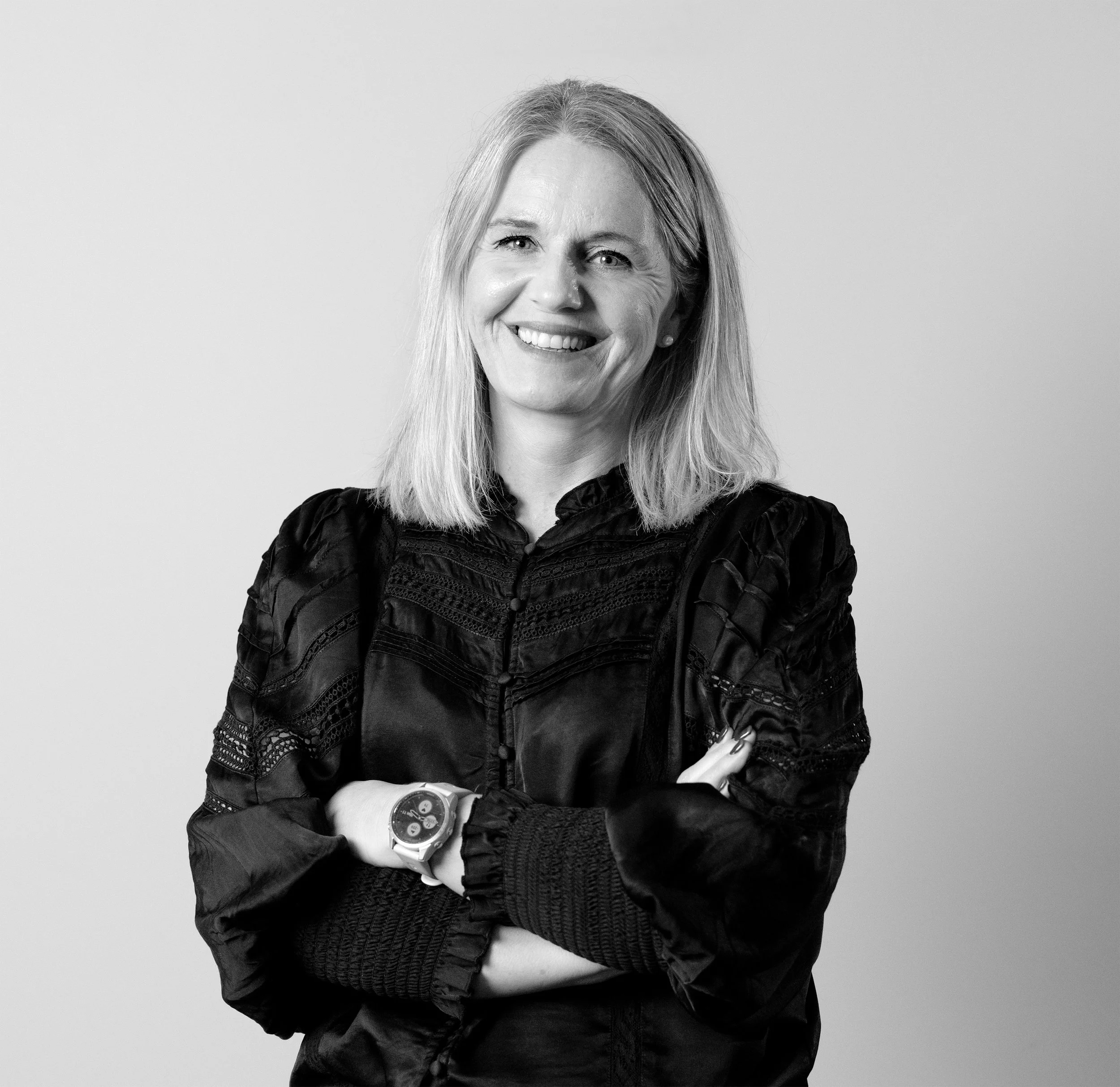 Et svart-hvitt portrett av en smilende kvinne med blondt hår, iført svart antrekk og armbåndsur, stående foran en lys bakgrunn.