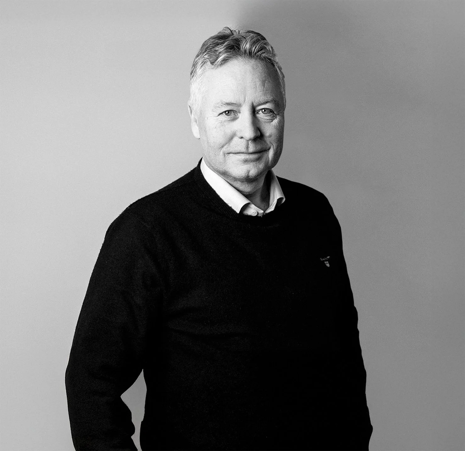 Et svart-hvitt bilde av en eldre mann med grått, kort hår, iført en mørk genser og en hvit skjorte, står foran en grå bakgrunn.