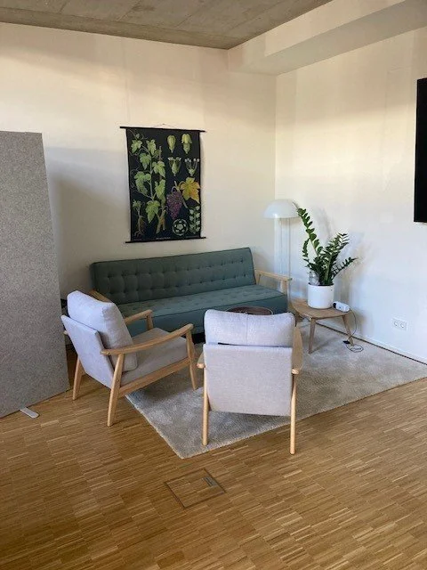 Gemütliches Wohnzimmer mit grünem Sofa, zwei weißen Armlehnenstühlen, einem kleinen Beistelltisch, Lampen und Pflanze vor weißer Wand mit Wandbild.