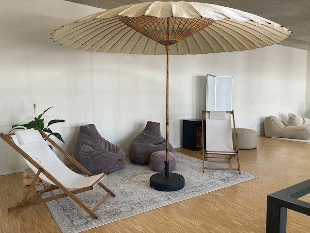 Gemütlicher Lounge-Bereich mit einem großen Sonnenschirm, einer Liege, zwei Sitzsäcken, einem Lounge-Chair, einem kleinen Fernseher, einem Hocker, und einem weißen Sofa auf einem Teppich.