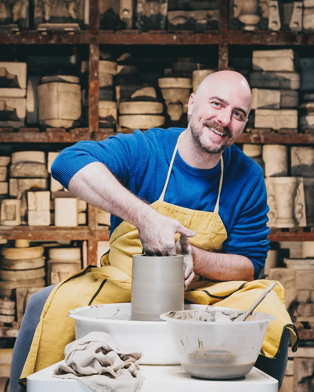 A man smiling while making pottery on a pottery wheel in a workshop with shelves of clay in the background.
