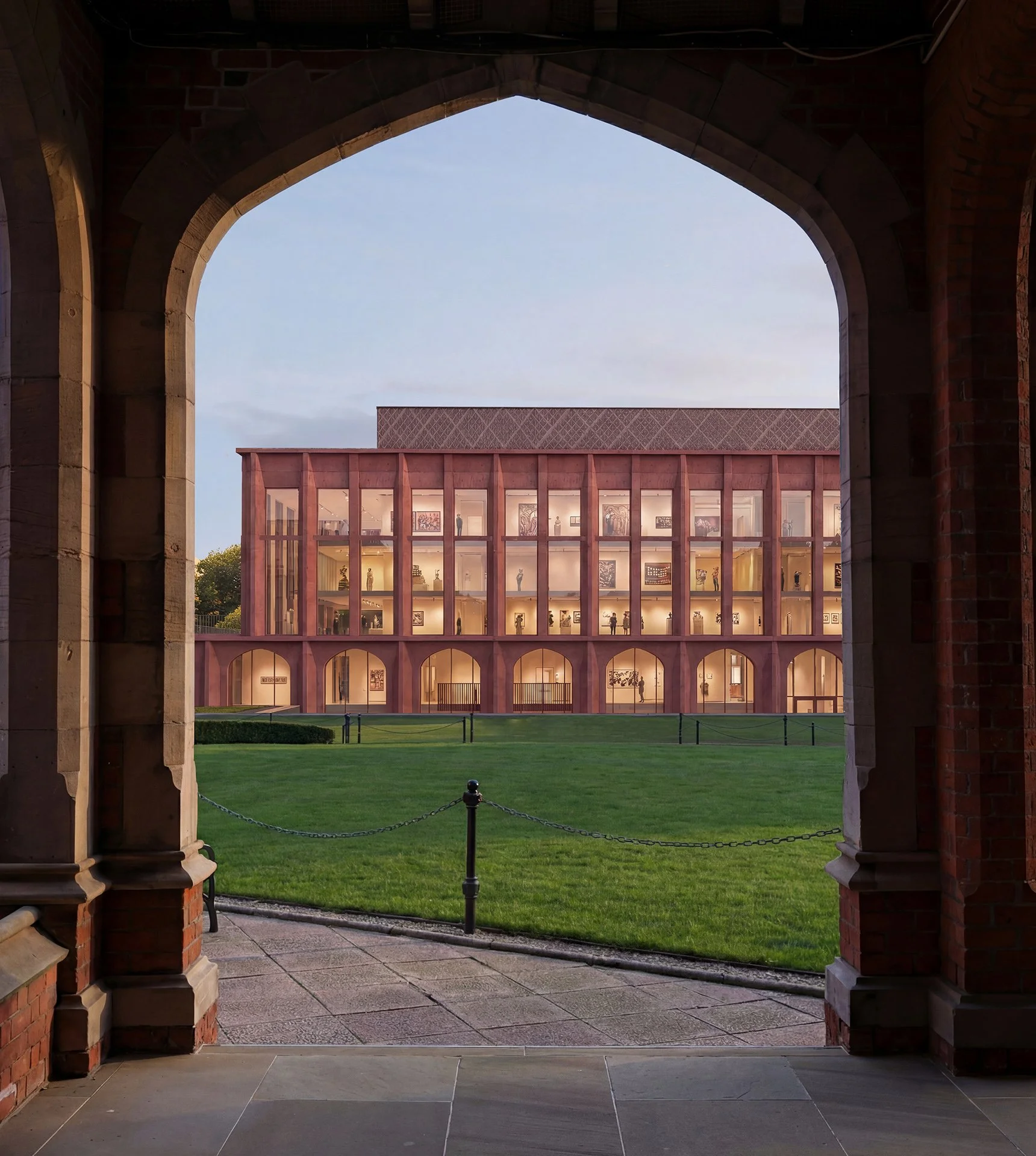 View of a modern, rectangular building with glass windows, framed by a brick and stone archway in the foreground. The building is illuminated from inside, displaying artwork or sculptures through the windows. The scene includes a well-maintained green lawn and a clear sky in the background.