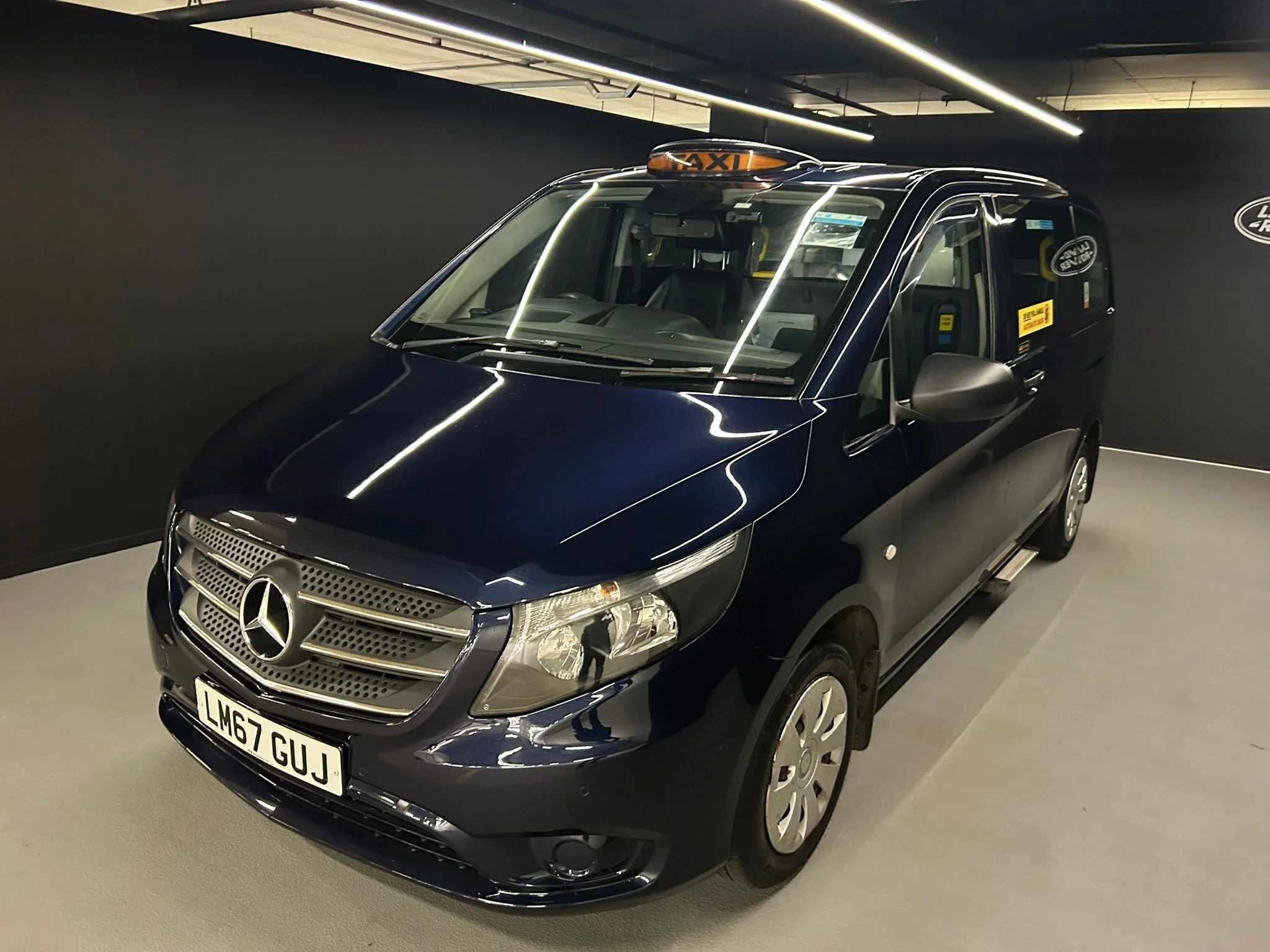 Dark blue Mercedes-Benz taxi with a roof sign reading 'TAXI', parked indoors. The vehicle has a UK license plate 'LM67 GUJ' and is surrounded by dark walls and ceiling with linear lighting.