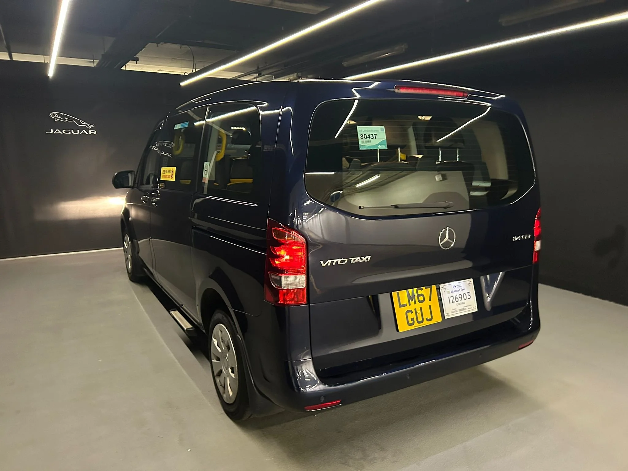 Black Mercedes-Benz Vito Taxi parked in an indoor showroom with a black wall featuring the Jaguar logo.
