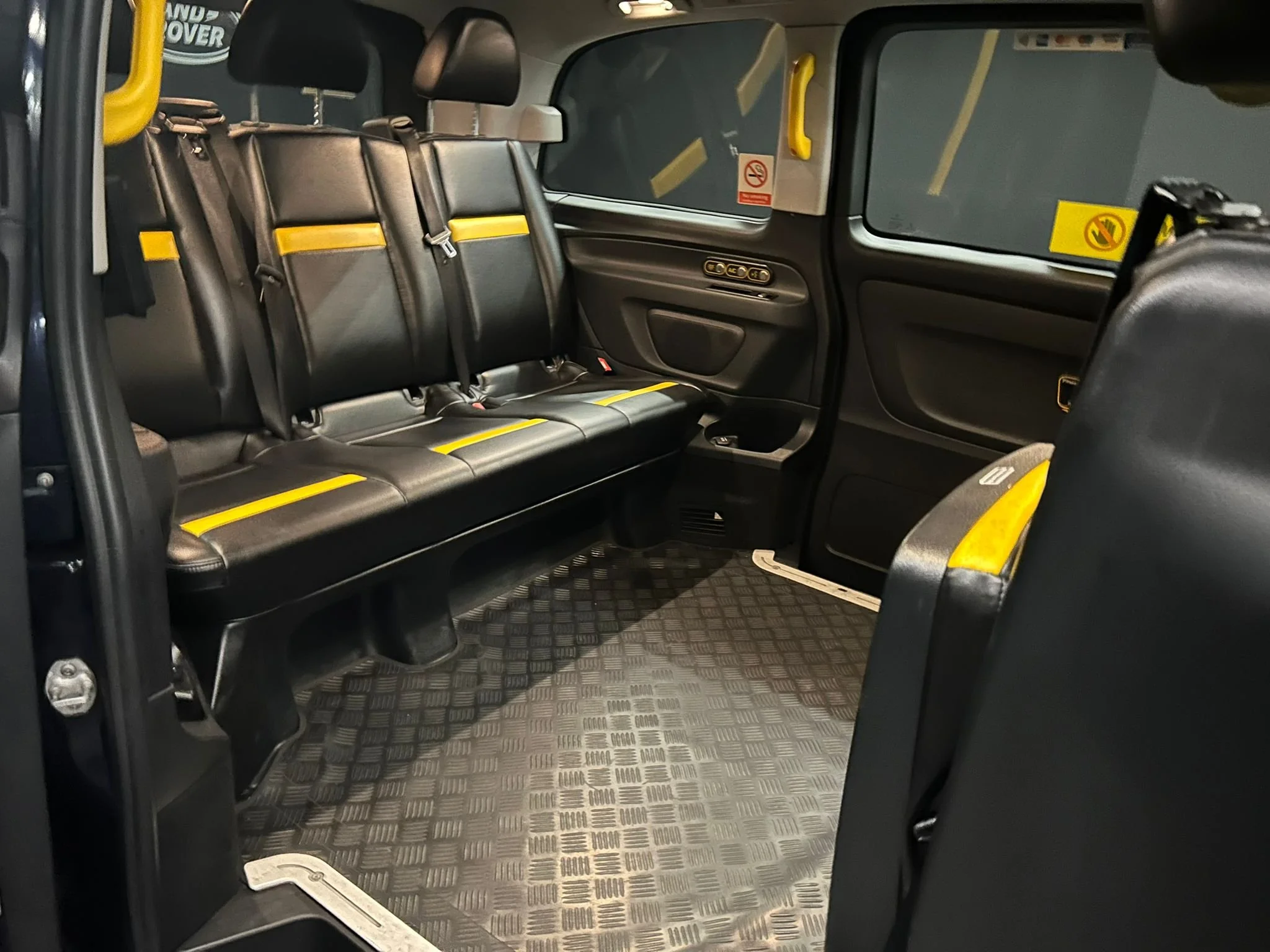 Interior of a black vehicle showing three black seats with yellow safety markings, windows with no smoking and no eating signs, and a checkered rubber floor.
