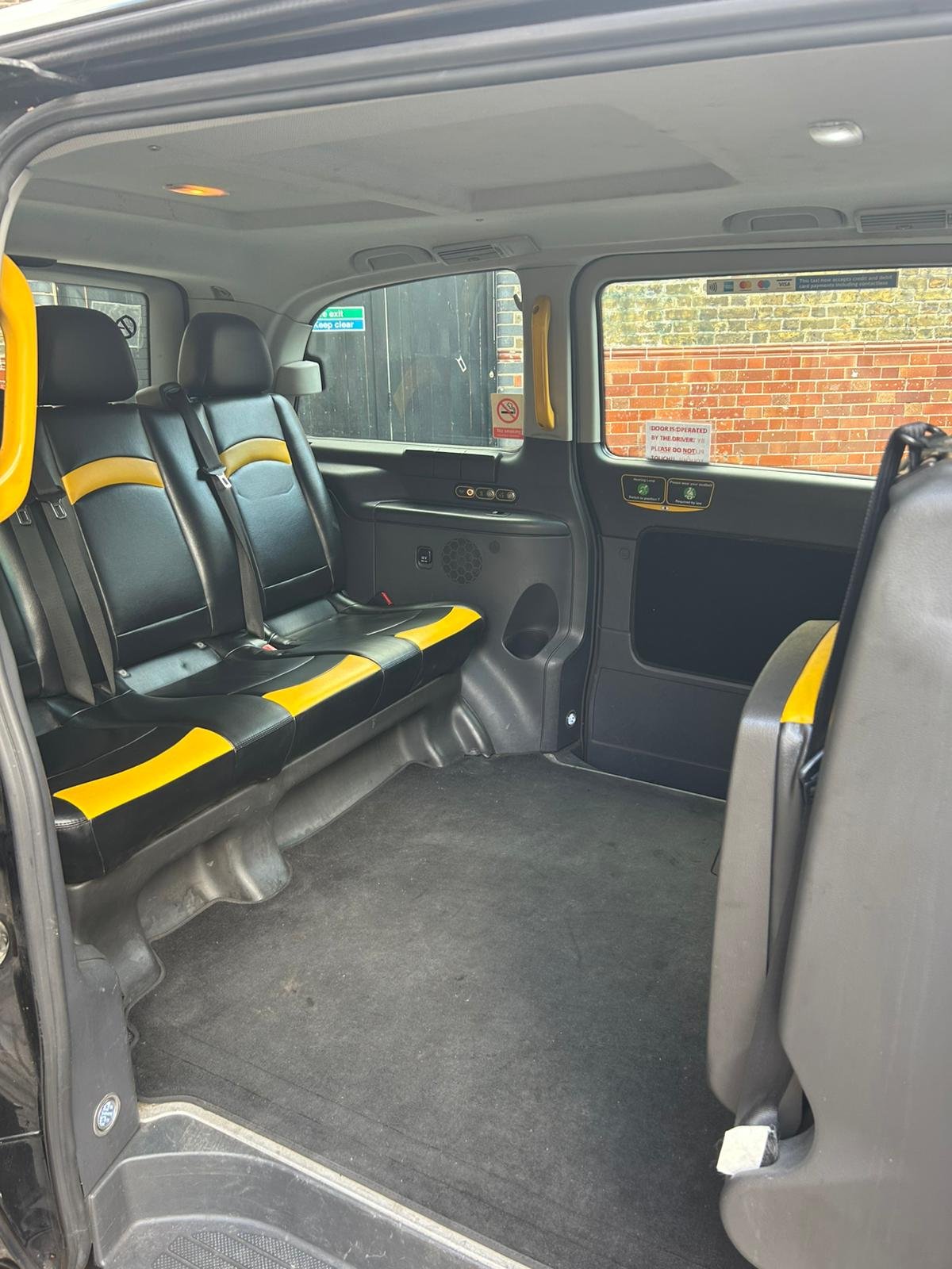 Interior of a passenger van with three black seats with yellow accents on the left side, a black floor, and a window showing a brick wall outside.