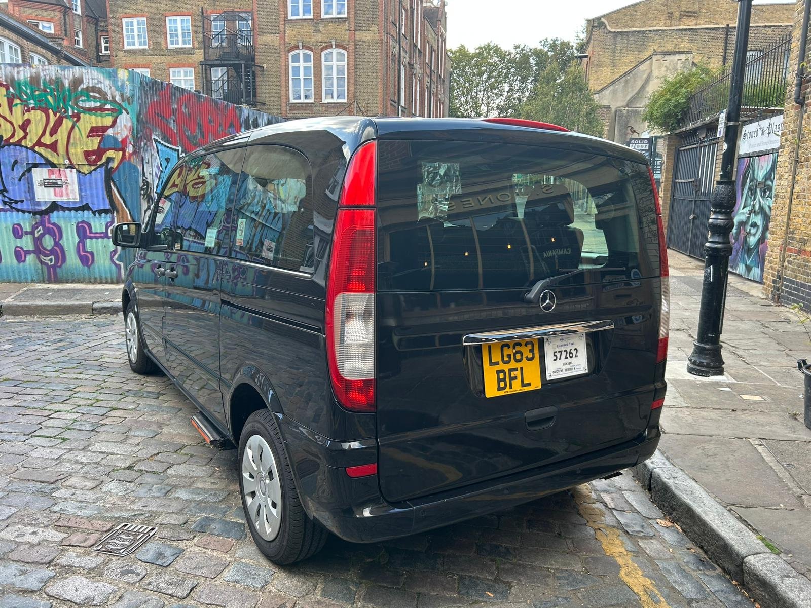 Black Mercedes-Benz van is parked on a cobblestone street with colorful graffiti on a corrugated metal fence in the background. There are buildings and trees visible behind the fence.