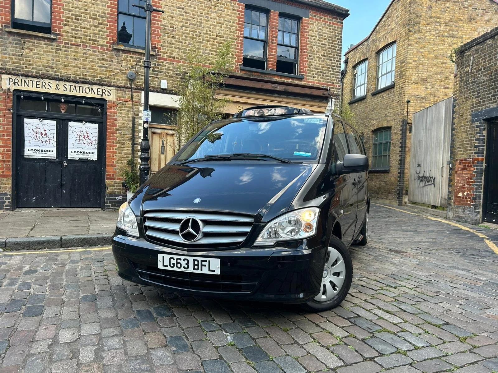 Black Mercedes-Benz taxi van parked on cobblestone street in front of old brick buildings with black doors and windows, one of which has 'Printers & Stationers' signage, and a wooden door with graffiti.