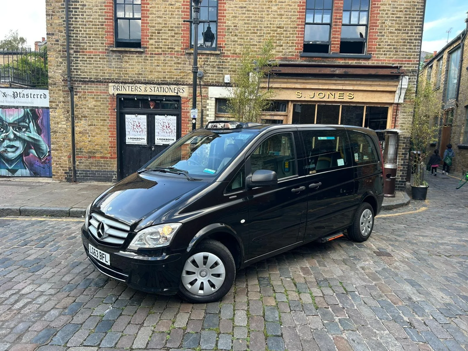 Black Mercedes-Benz taxi parked on cobblestone street in front of brick building with signs 'Prints & Stationers' and 'S. Jones'.
