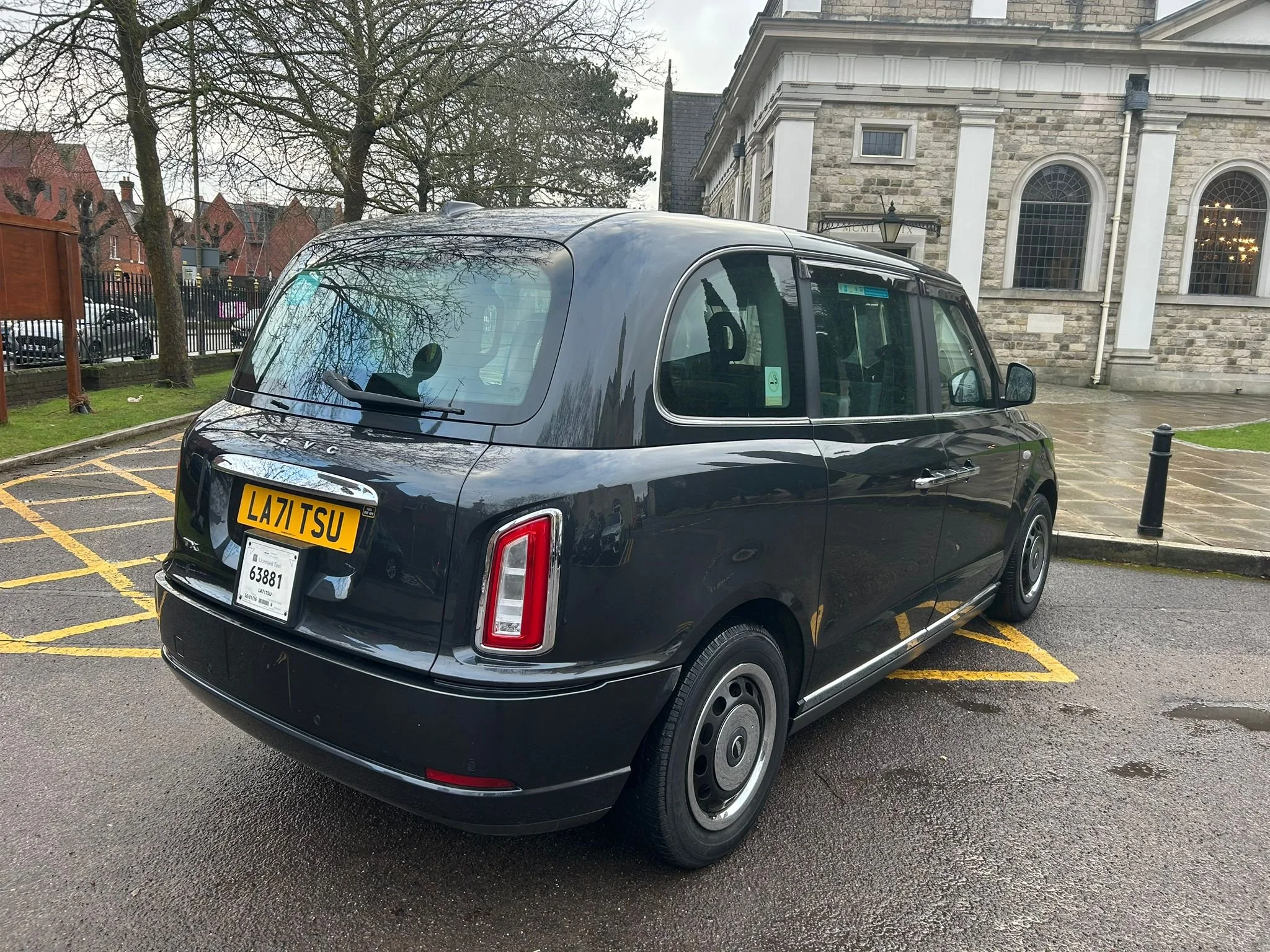A black London taxi cab parked in a designated parking spot on a wet pavement, with a historic stone building and leafless trees in the background.