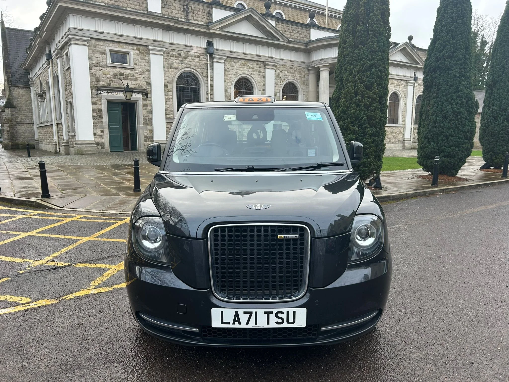 Front view of a black London taxi cab parked on wet pavement in front of a stone building with columns and tall trees.