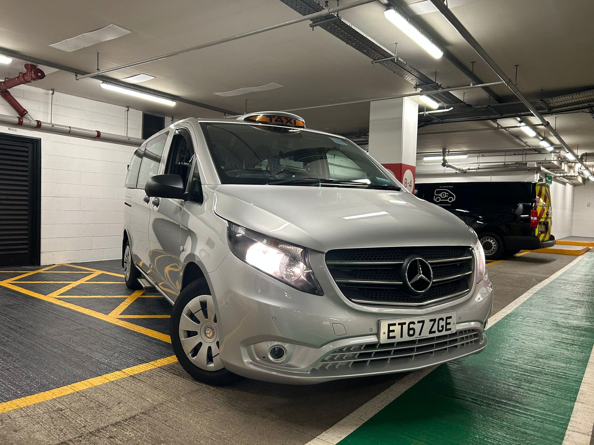 A silver Mercedes-Benz taxi van parked in an indoor parking garage, positioned in a designated accessible parking space with yellow stripes.