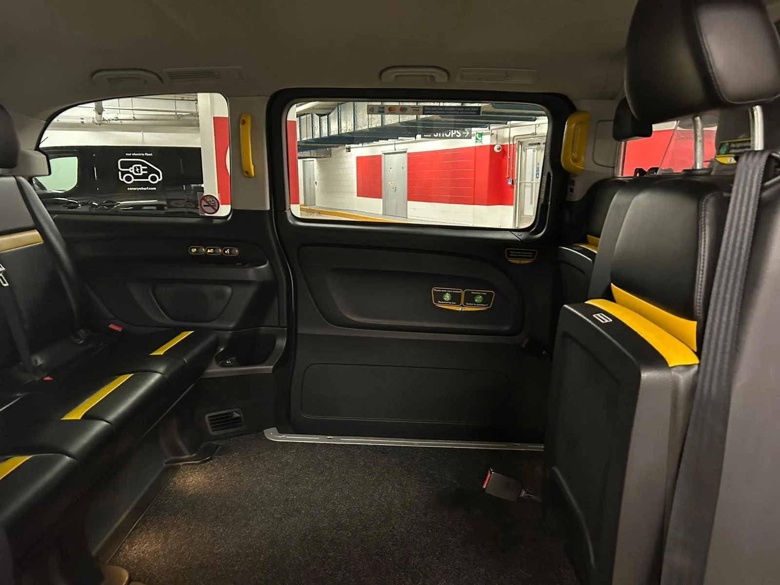 Interior of a vehicle, likely a shuttle or bus, with black seats and yellow safety strips. The image shows the back seats, a window, and a door to the parking garage.