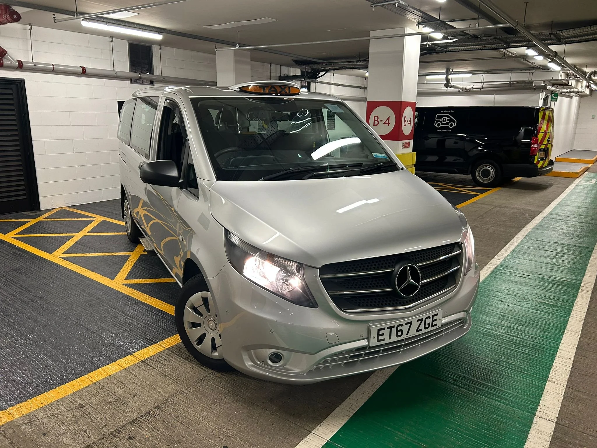 Silver Mercedes-Benz taxi van parked in a designated parking space in an indoor parking garage, with a black van parked behind it.