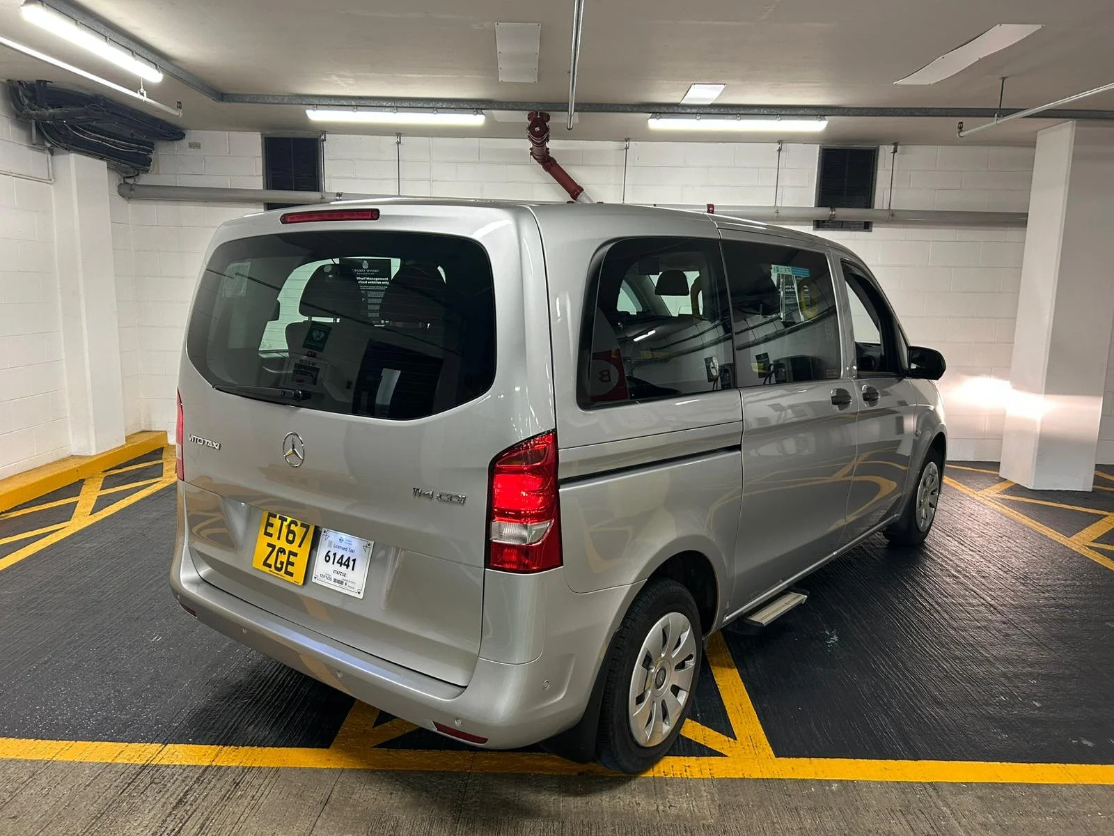 A silver Mercedes-Benz Vito taxi van parked in a designated parking space in an indoor parking garage, with yellow markings on the floor and white brick walls.