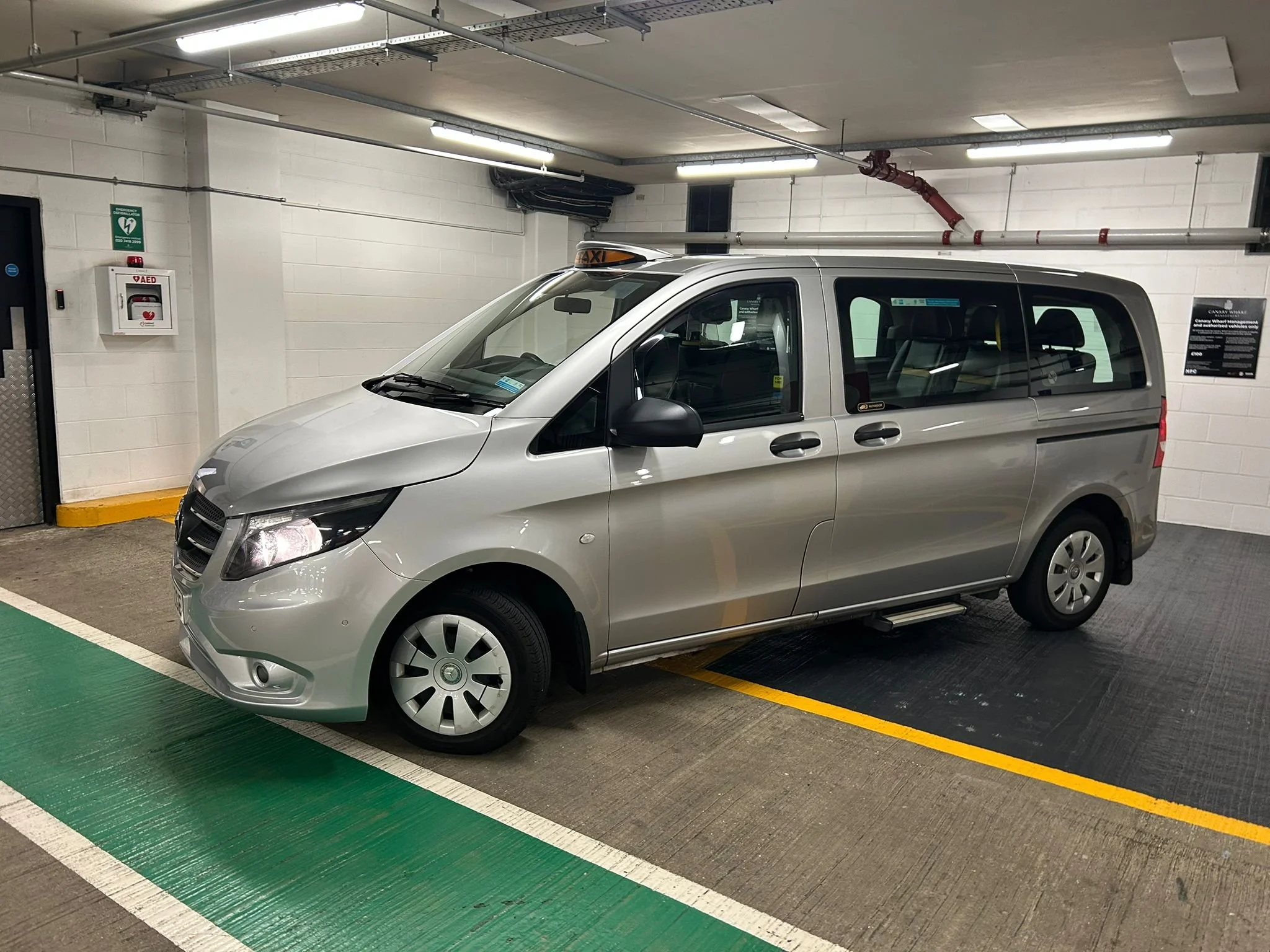 Silver taxi van parked in an indoor parking garage with fire safety equipment in the background.
