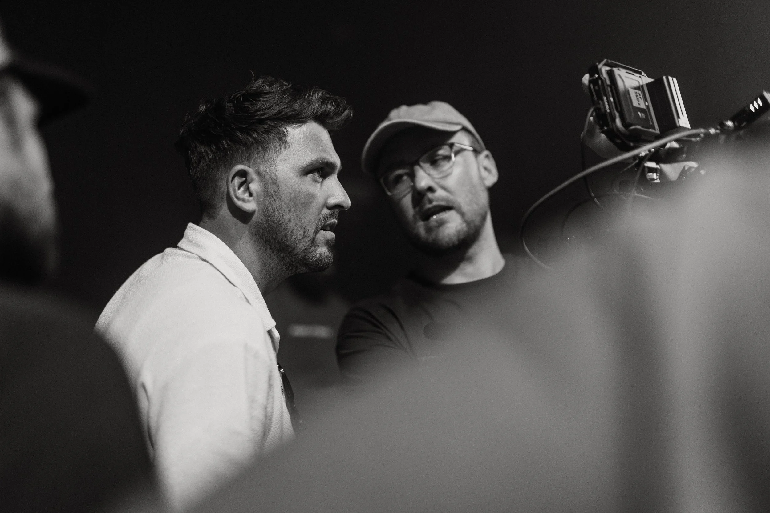 Two men are engaged in a conversation or interview; one man with short hair and stubble is facing slightly to the left, while the other man with glasses and a cap is turned towards him. A camera is visible recording or photographing them.