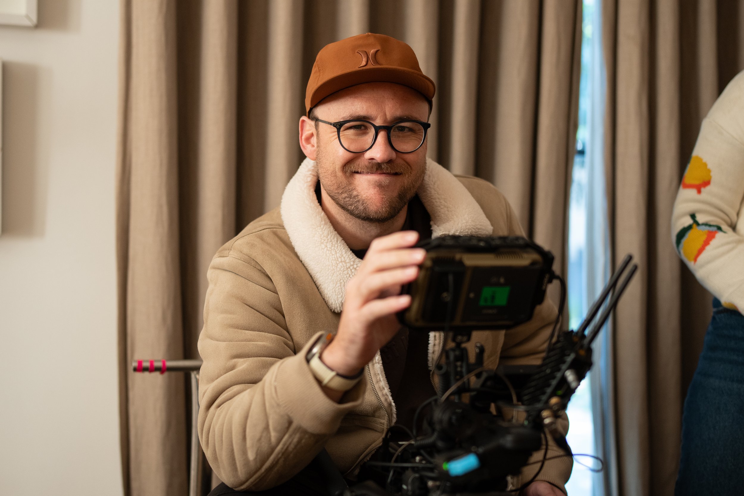 Man with glasses, wearing a brown cap and beige jacket, working with a camera rig.