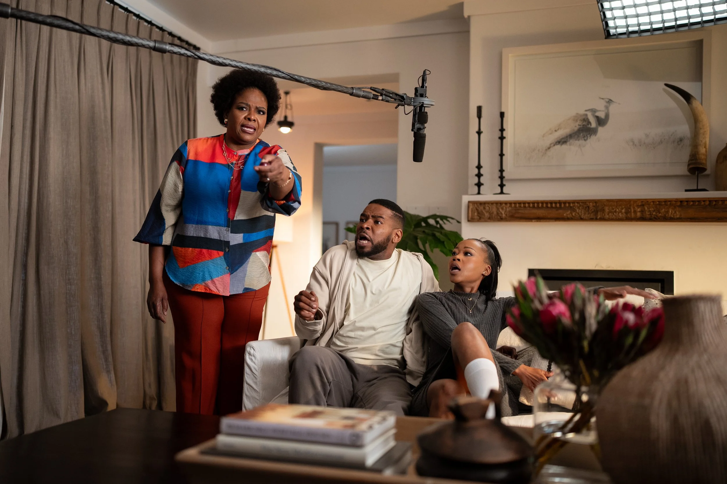 Three people in a living room, appearing surprised or confused, with one woman standing and two seated individuals looking up. There is a microphone overhead, and the room has artwork and curtains.