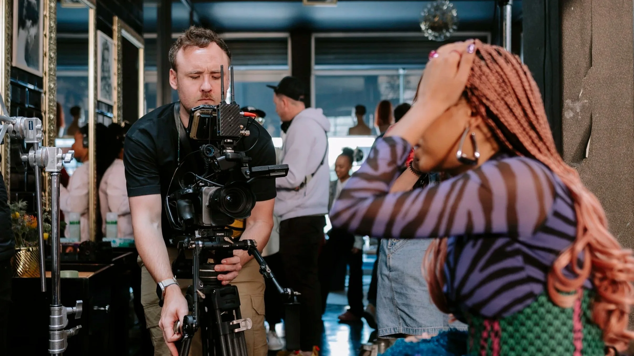 A man operating a professional video camera on a tripod at an indoor event with a woman in a striped jacket and pink braids in the foreground, and several people in the background.