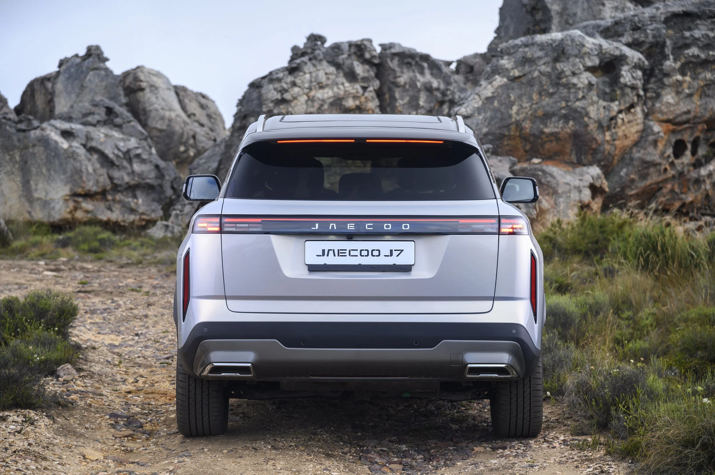 A silver SUV parked on a dirt path surrounded by rocky hills and sparse green vegetation.