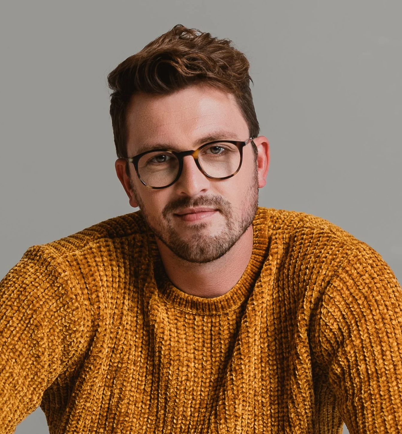 A man with short brown hair, glasses, and a beard is wearing a mustard yellow textured sweater and looking at the camera against a plain gray background.