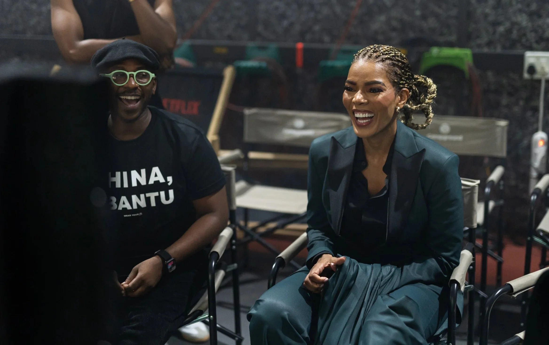 Two women sitting and laughing at a table in a restaurant or café, with one wearing glasses and a black shirt and the other with braided hair and a dark blazer, in a lively indoor setting.