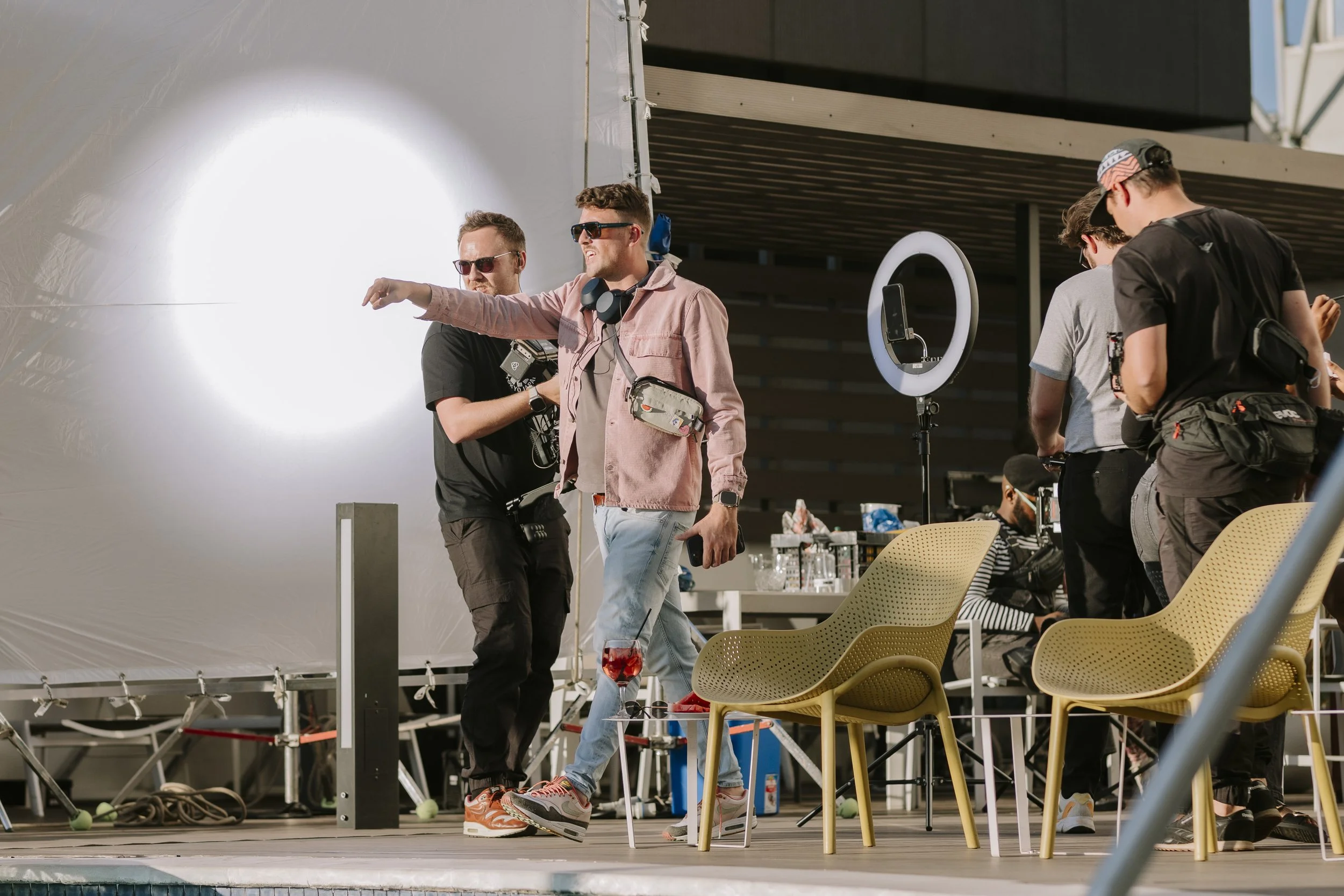 A group of people on a film set, with crew members and equipment, including lights, chairs, and a ring light, near a swimming pool during daytime.