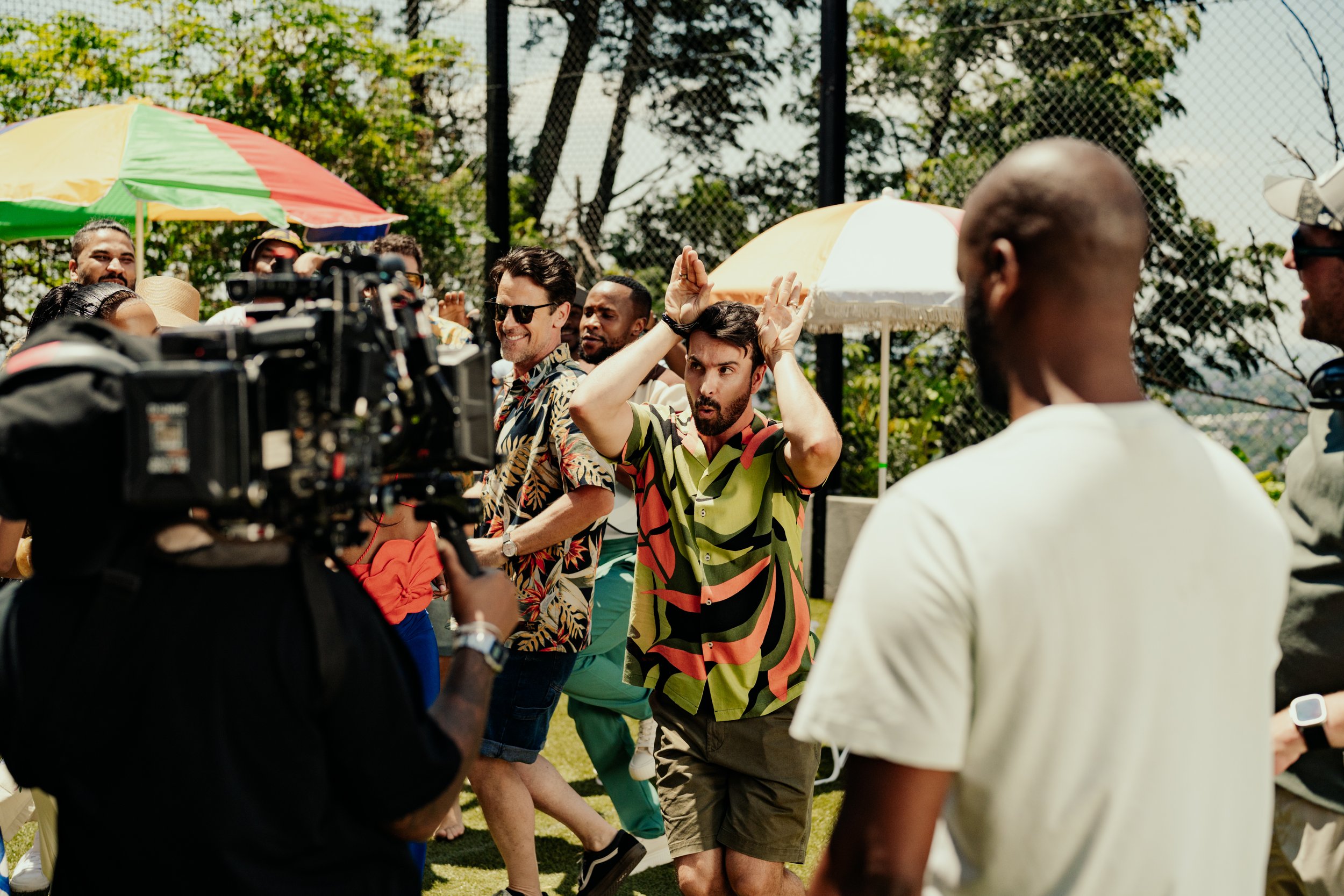 Group of people at an outdoor event, some dancing and enjoying themselves, with colorful umbrellas and trees in the background.