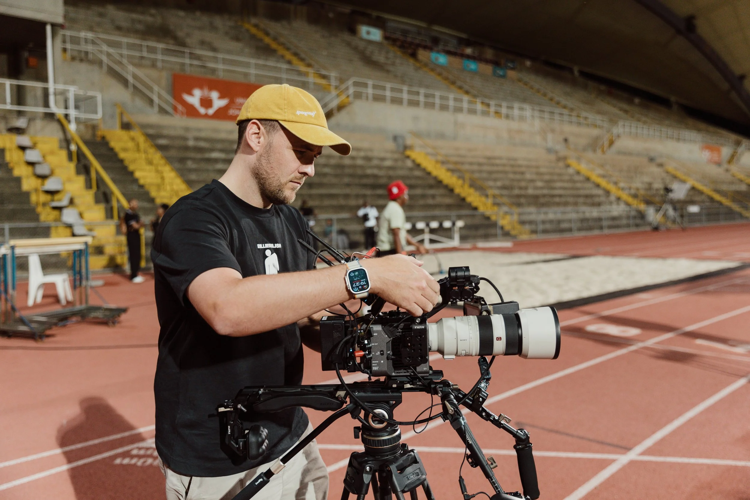 A man with a yellow cap and black shirt operating a professional video camera on a tripod at a sports stadium, with empty bleachers and track field in the background.