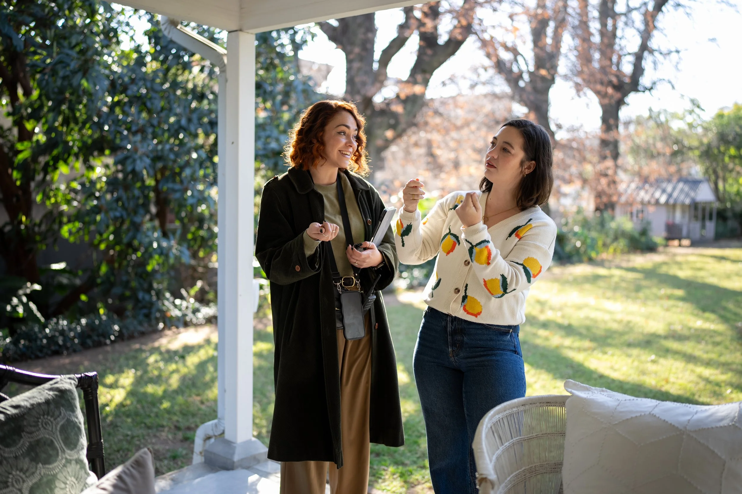 Two women are talking outside under a porch in a yard with trees and a small house in the background. One woman has curly red hair, is smiling, and holds a smartphone. The other woman has dark hair, is wearing a patterned sweater, and is gesturing wi