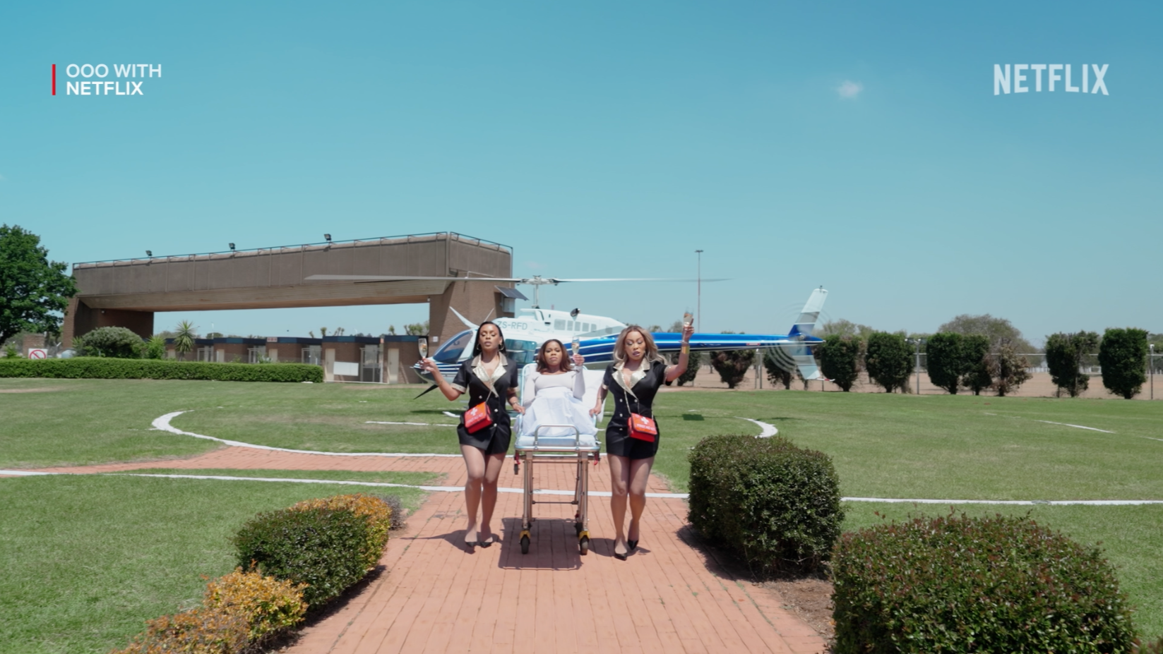 Three women dressed as airline staff escorting a person on a stretcher across an airport tarmac, with a helicopter in the background on a clear day.