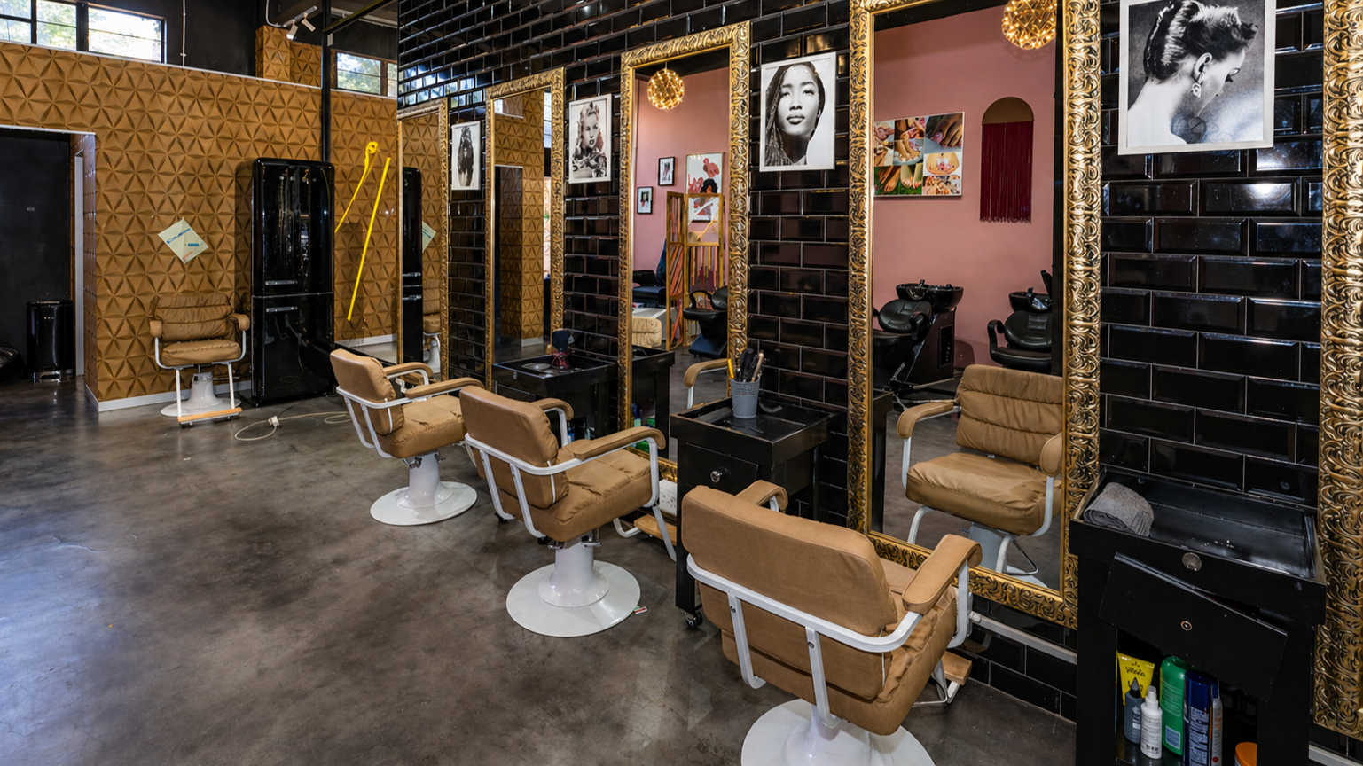 A modern hair salon with multiple styling stations. Each station has a mirror with ornate gold frames, a beige salon chair, and various styling tools. The space features a mix of black and gold decor, with brick walls, geometric wall textures, and framed black-and-white photographs.