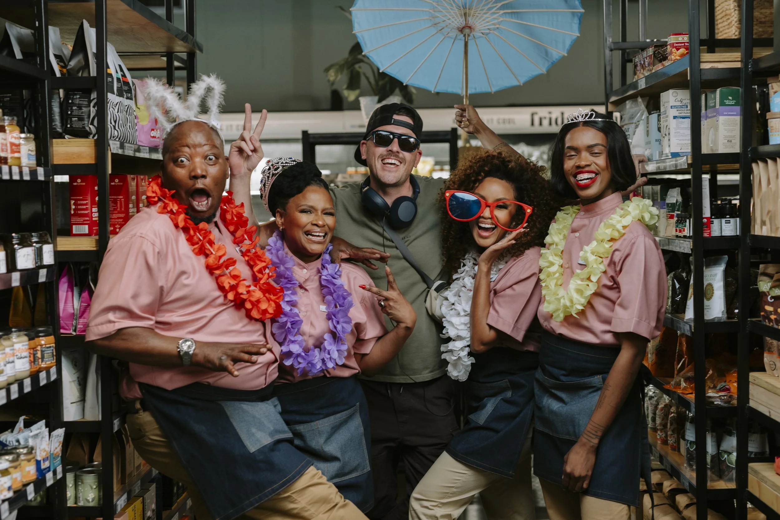 Group of six diverse people in a grocery store aisle celebrating, wearing colorful leis, posing playfully, with one person holding a blue umbrella.