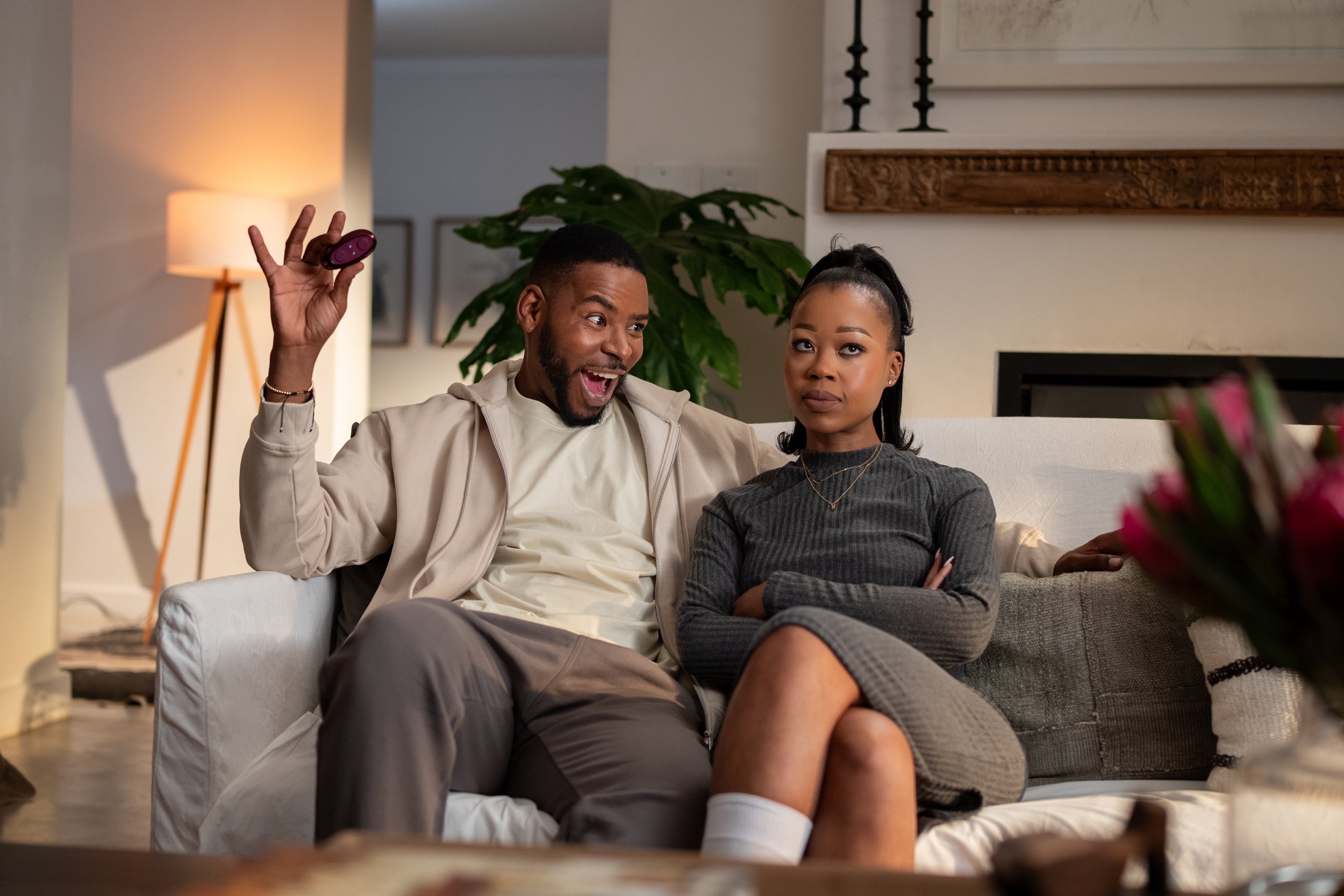 A man and woman sitting on a gray sofa in a living room. The man is holding a purple remote control and smiling excitedly, while the woman has a serious expression with crossed arms. There is a large green plant behind them, a framed picture on the w