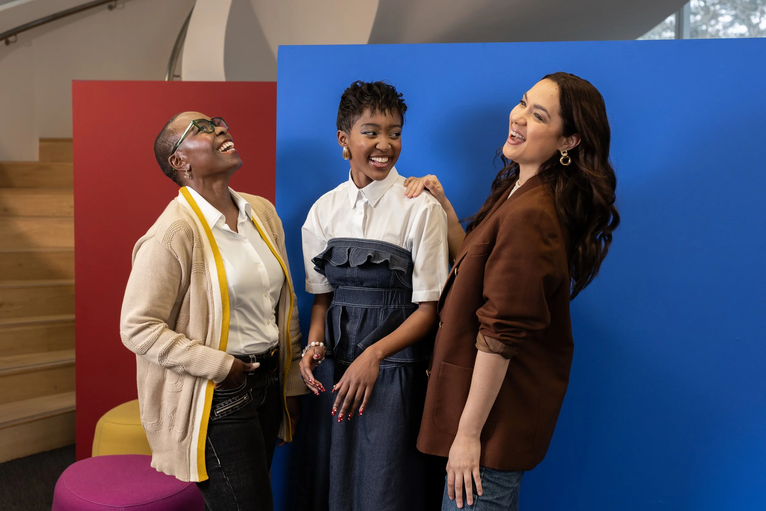 Three women smiling and interacting in front of a blue and red backdrop. One woman has short hair, glasses, and a beige cardigan, another has short hair and a white shirt, and the third has long hair and a brown blazer.