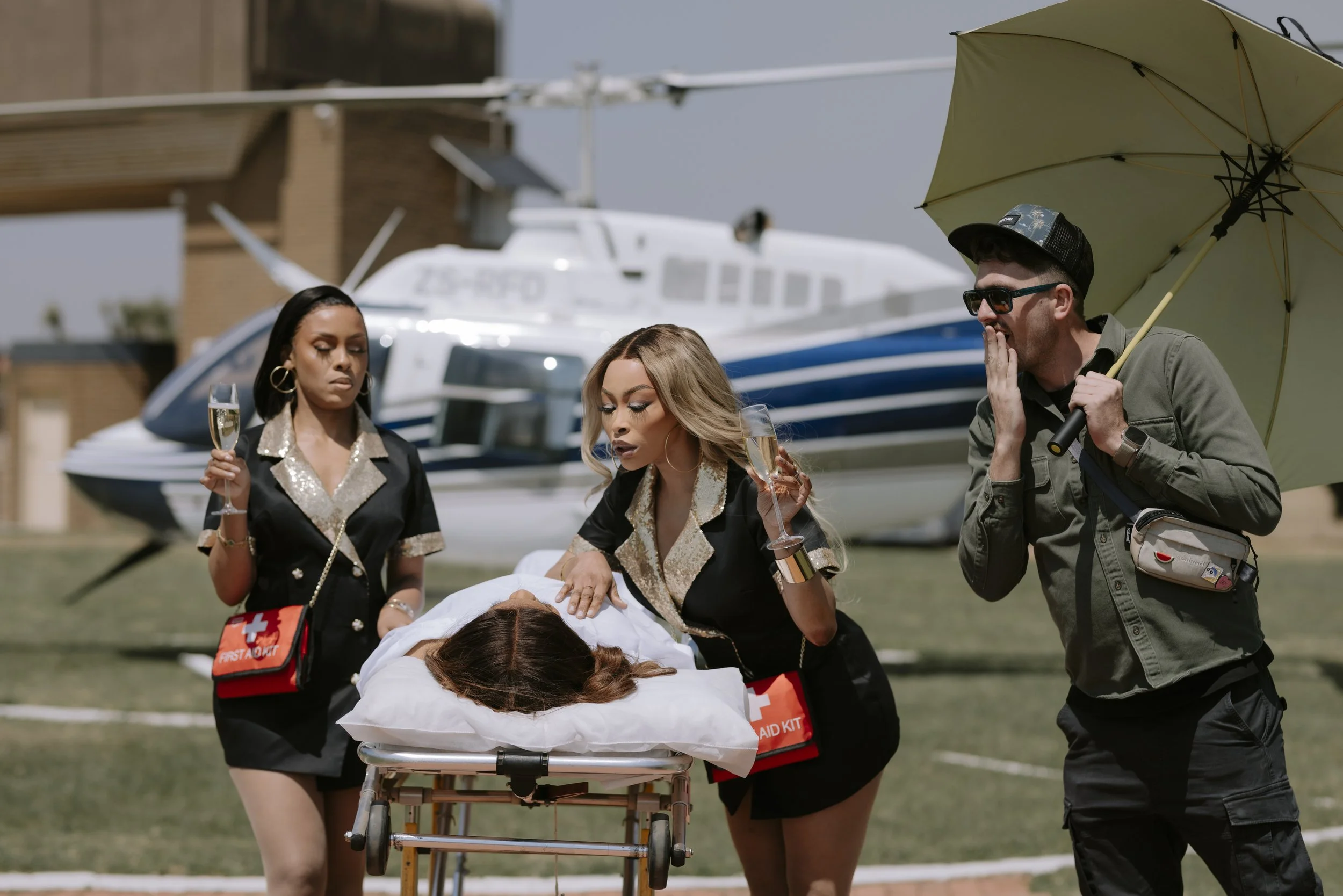 People providing emergency medical care to a person on a stretcher with medical supplies, a helicopter, and a person with an umbrella in the background.