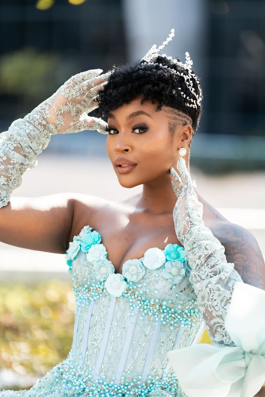 A woman with short curly hair styled in a fade with curls on top, wearing a strapless light blue dress decorated with flowers and beads, white lace gloves, earrings, and makeup, posing outdoors with a blurred background.
