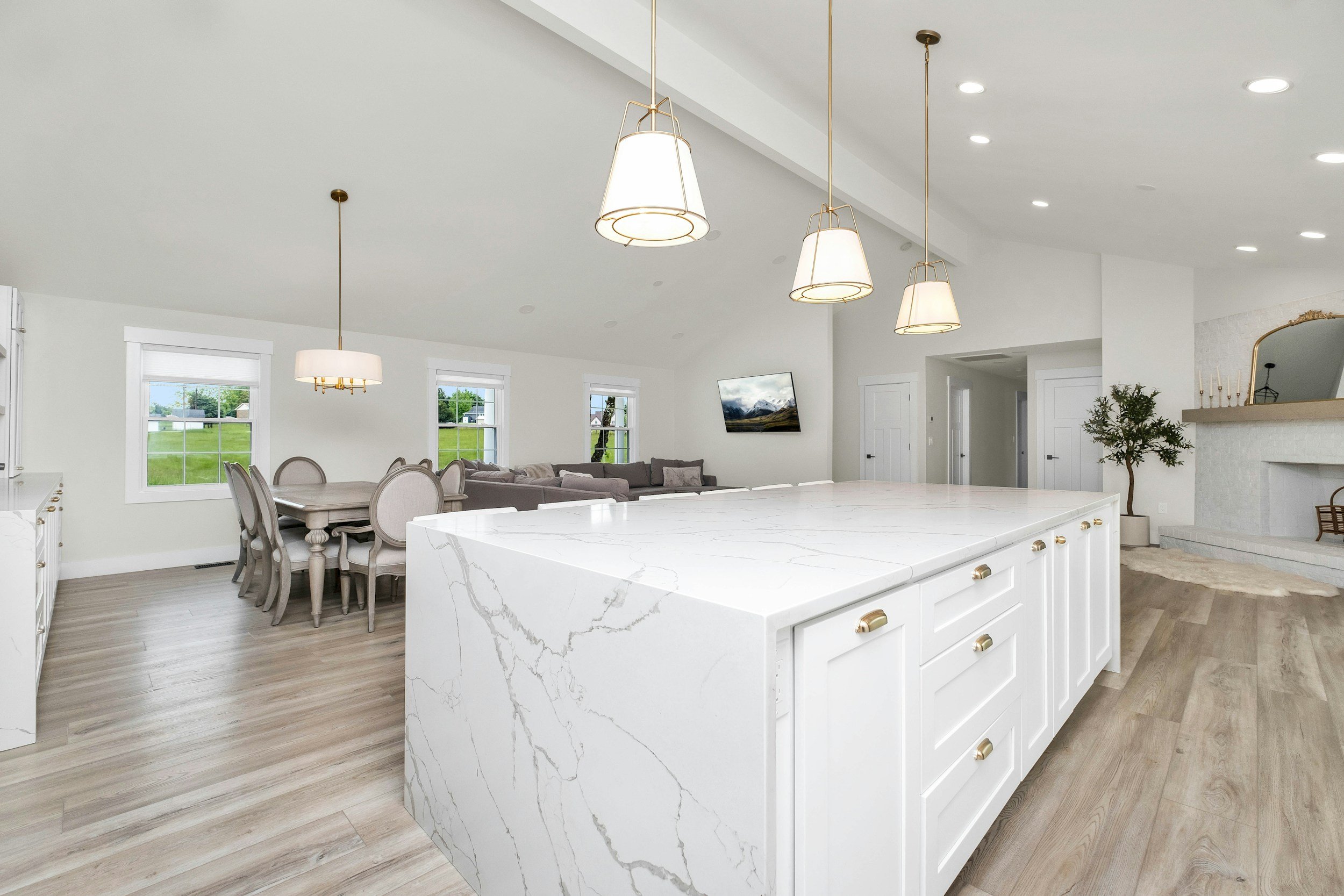 Bright, modern open-concept kitchen and dining area with white walls, a marble island, hardwood floors, large windows, and stylish pendant and chandelier lighting.