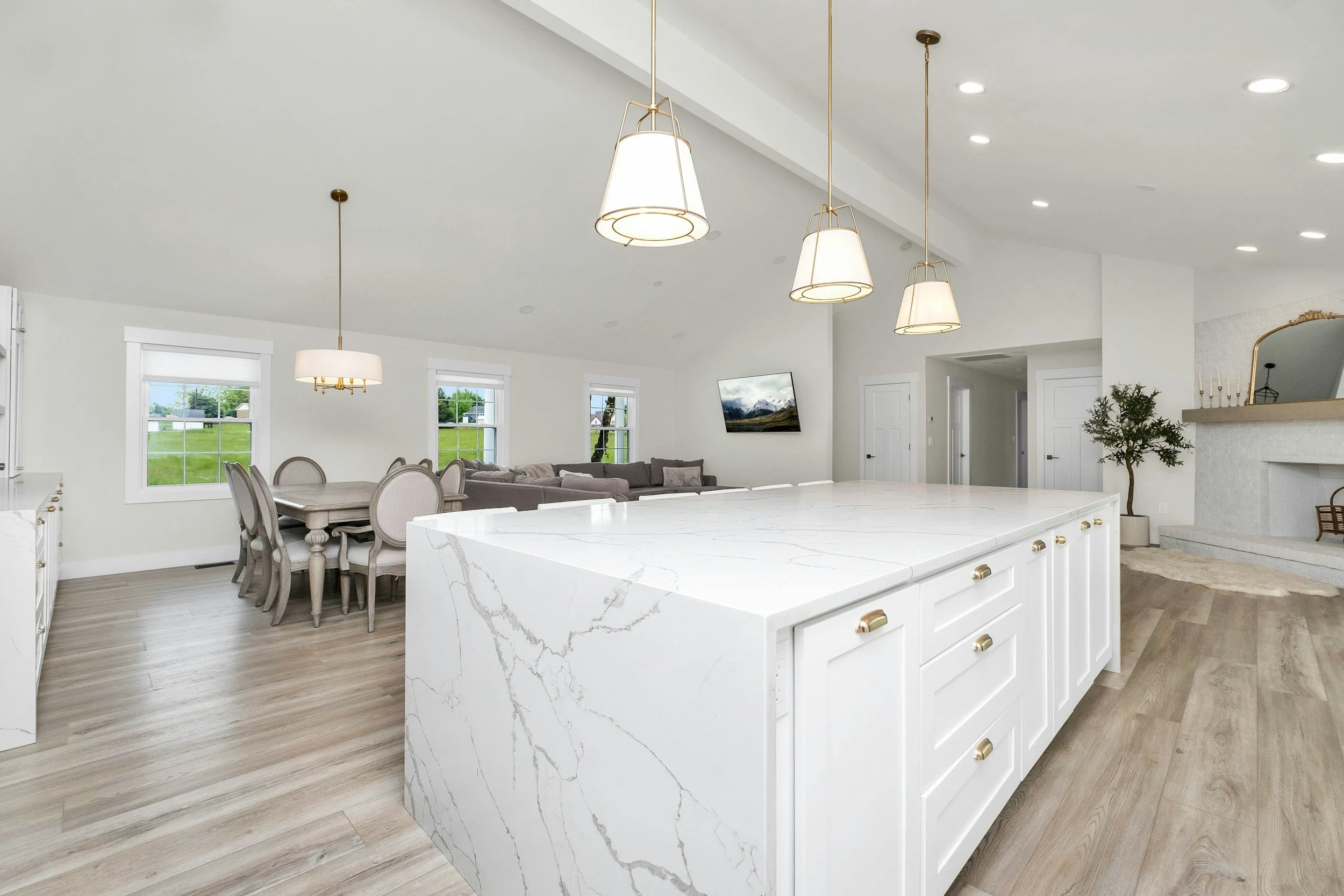 Modern open-concept kitchen with white marble island, light wood flooring, and hanging pendant lights. Adjacent dining area with a table and chairs, and living space with a couch and wall-mounted TV.
