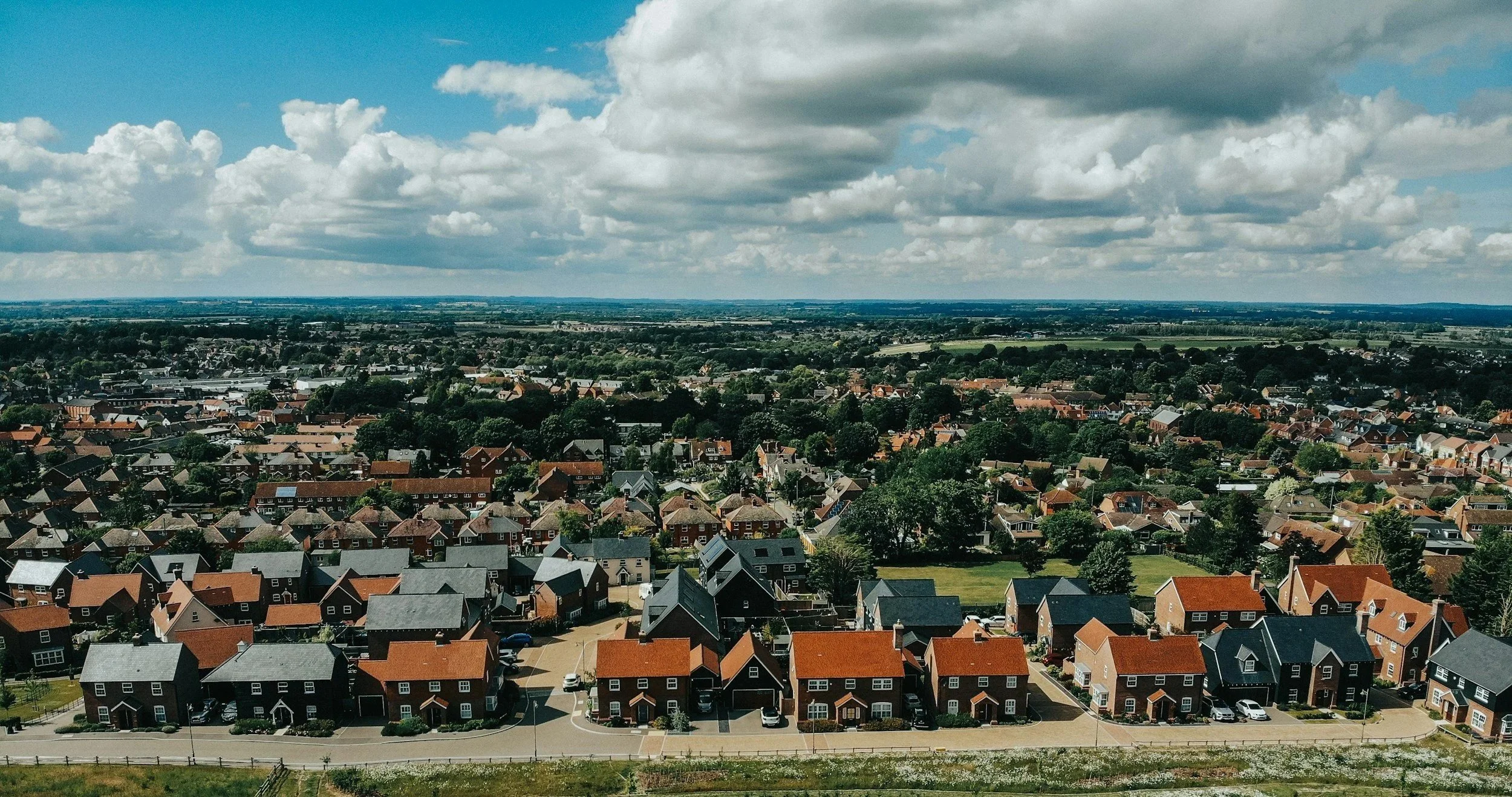 Aerial view of a suburban neighborhood with rows of houses, trees, and streets under a partly cloudy sky.